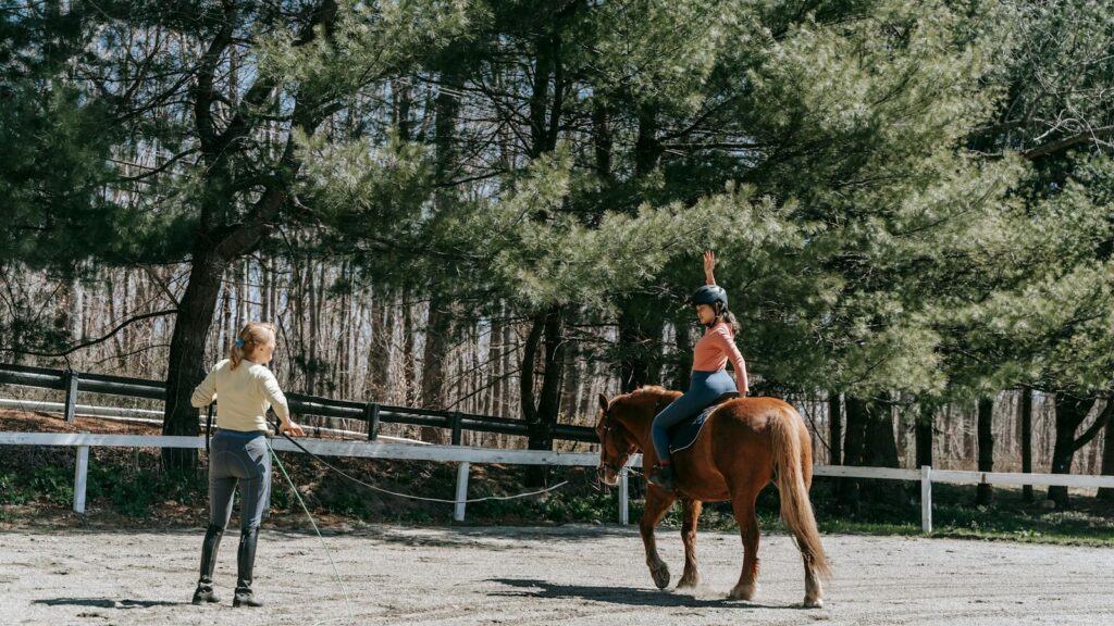 Woman instructing a child horseback riding under sunny forest trees.