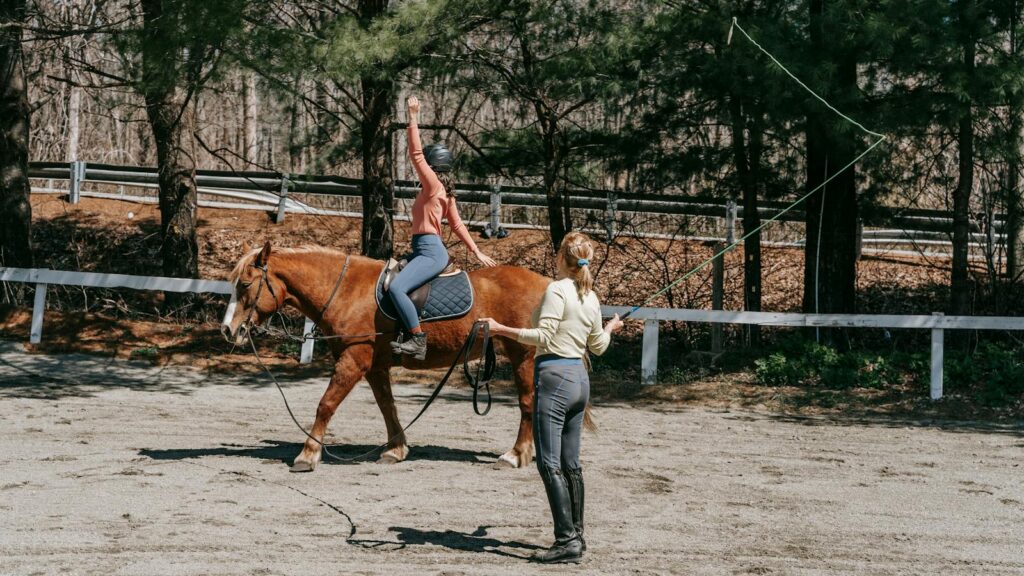 Two women practicing horseback riding in a sunny outdoor arena.