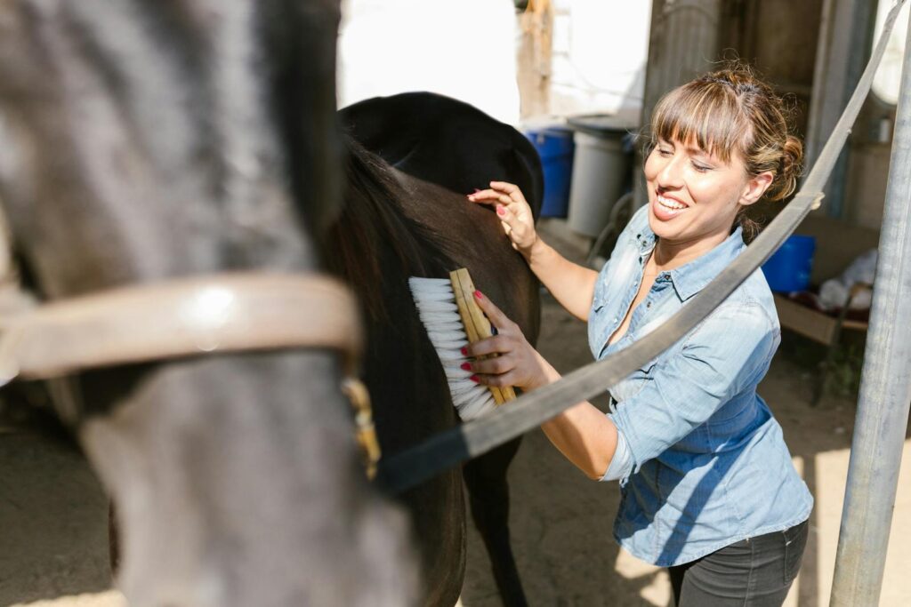 Smiling woman brushing a horse in a sunny outdoor stable, emphasizing care and connection.