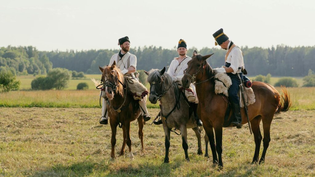Three men in historical uniforms riding horses in a open field with a forest backdrop.